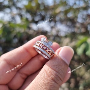 May include: A silver-toned ring featuring a top section shaped like a mouse head with two circular ears. The band is adorned with a row of small clear stones and a central row of red heart-shaped stones. The text Meerajewelsart is visible.