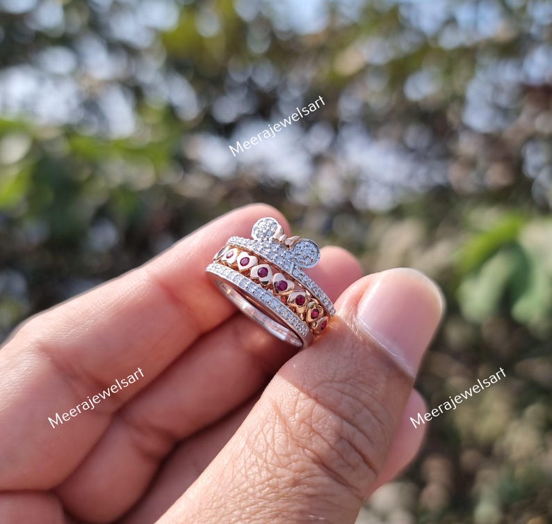 May include: A silver-toned ring featuring a top section shaped like a mouse head with two circular ears. The band is adorned with a row of small clear stones and a central row of red heart-shaped stones. The text Meerajewelsart is visible.