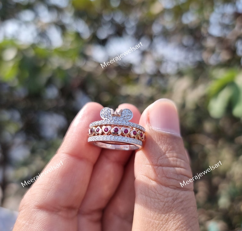 May include: A multi-band ring featuring a top section shaped like a mouse head with white stones, a middle band with a row of small red circular stones set within heart-shaped metal frames, and a bottom band encrusted with small white stones.