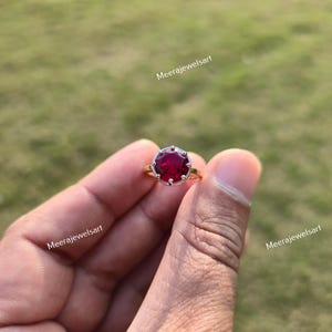 Può includere: Un anello d'oro con una grande gemma rotonda di colore rosso intenso. La gemma è incastonata in una lunetta color argento con un bordo smerlato. L'anello è tenuto in mano su uno sfondo verde sfocato. Il testo "Meerajewelsart" è visibile.