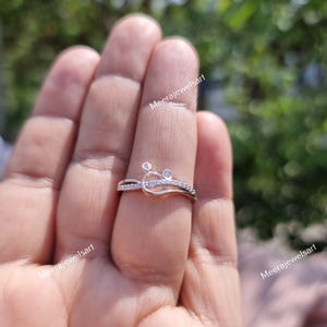 May include: A rose gold ring with a unique design featuring small, clear gemstones. The ring has a delicate, intertwined pattern with a small, circular accent. The ring is shown on a finger.