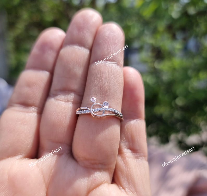 May include: A rose gold ring with a unique design featuring small, clear gemstones. The ring has a delicate, intertwined pattern with a small, circular accent. The ring is shown on a finger.