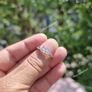 May include: A delicate silver ring with a heart-shaped design, accented with small, sparkling stones. The ring features a curved band and is set against a blurred green background. The ring is a piece of jewelry.