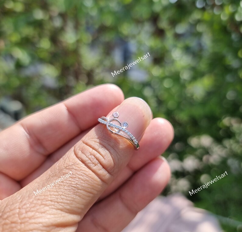May include: A delicate silver ring with a heart-shaped design, accented with small, sparkling stones. The ring features a curved band and is set against a blurred green background. The ring is a piece of jewelry.