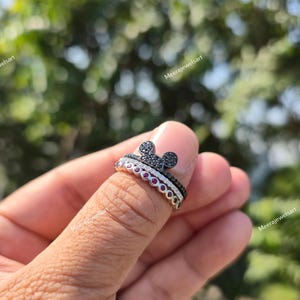 K&ouml;nnte beinhalten: Ein stapelbares Ring-Set, bestehend aus einem oberen Ring mit einem mit schwarzen Steinen besetzten Mausohren-Motiv und einem unteren Ring, der mit einer Reihe runder roter Steine besetzt ist, gefertigt aus poliertem Metall.