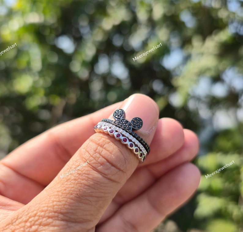 K&ouml;nnte beinhalten: Ein stapelbares Ring-Set, bestehend aus einem oberen Ring mit einem mit schwarzen Steinen besetzten Mausohren-Motiv und einem unteren Ring, der mit einer Reihe runder roter Steine besetzt ist, gefertigt aus poliertem Metall.