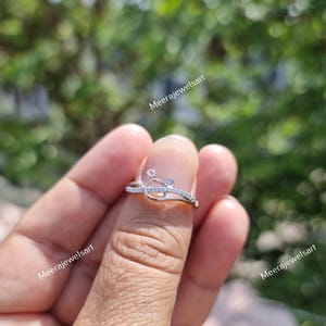May include: A silver ring with a delicate design featuring small, clear gemstones. The ring has a unique shape with a curved band and is adorned with multiple small, sparkling stones. The ring is shown on a finger.
