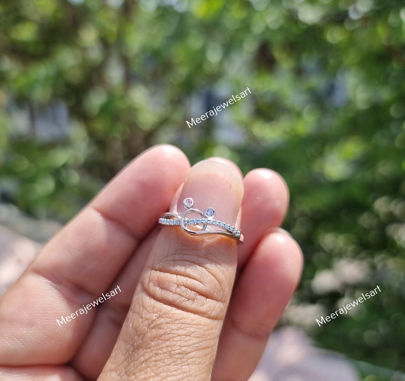 May include: A silver ring with a delicate design featuring small, clear gemstones. The ring has a unique shape with a curved band and is adorned with multiple small, sparkling stones. The ring is shown on a finger.