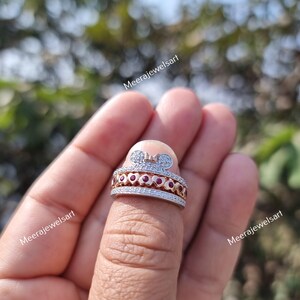 May include: A two-tone ring featuring a silver-colored band with pav&eacute; stones and a rose gold-colored center band set with small red stones. The top of the ring displays a stylized mouse ear silhouette with a small bow detail and pav&eacute; stones.