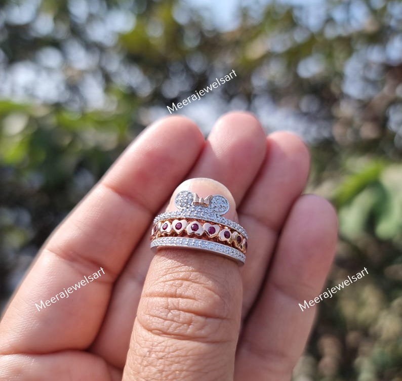 May include: A two-tone ring featuring a silver-colored band with pav&eacute; stones and a rose gold-colored center band set with small red stones. The top of the ring displays a stylized mouse ear silhouette with a small bow detail and pav&eacute; stones.