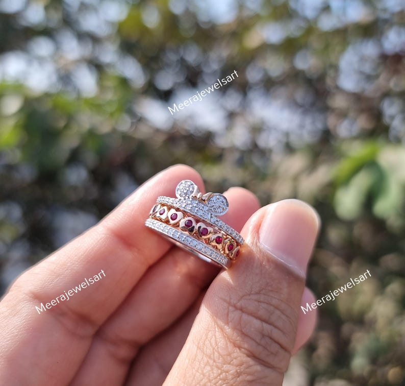 May include: A silver-toned ring featuring a band with a row of small clear stones, a central row of red heart-shaped stones, and a top section shaped like a mouse head with clear stones. The text Meerajewelsart is visible on the image.