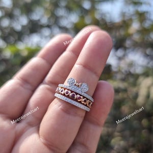 May include: A multi-band ring featuring a top band with a bow shape encrusted with small clear stones, a middle band with a row of small red heart-shaped stones, and a bottom band with a row of small clear stones, all set in a metallic finish.