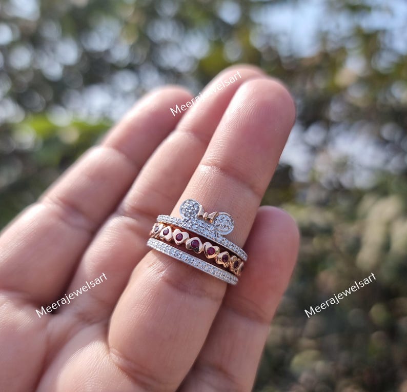 May include: A multi-band ring featuring a top band with a bow shape encrusted with small clear stones, a middle band with a row of small red heart-shaped stones, and a bottom band with a row of small clear stones, all set in a metallic finish.