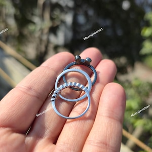 K&ouml;nnte beinhalten: Ein Set aus drei gestapelten Ringen, bestehend aus einem dunklen Metallring mit einer kleinen Schleifenverzierung, einem silberfarbenen Ring mit einer Reihe kleiner klarer Steine und einem schlichten silberfarbenen Ring.