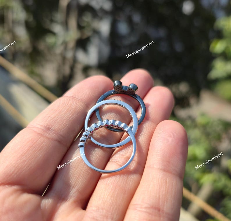 K&ouml;nnte beinhalten: Ein Set aus drei gestapelten Ringen, bestehend aus einem dunklen Metallring mit einer kleinen Schleifenverzierung, einem silberfarbenen Ring mit einer Reihe kleiner klarer Steine und einem schlichten silberfarbenen Ring.