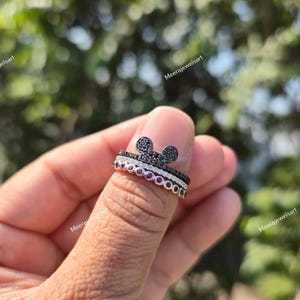 K&ouml;nnte beinhalten: Ein stapelbares Ring-Set bestehend aus einem oberen Ring mit einer mit schwarzen Steinen besetzten Mausohr-Silhouette, einem mittleren Ring mit klaren Pav&eacute;-Steinen und einem unteren Ring mit einer Reihe runder roter Steine.