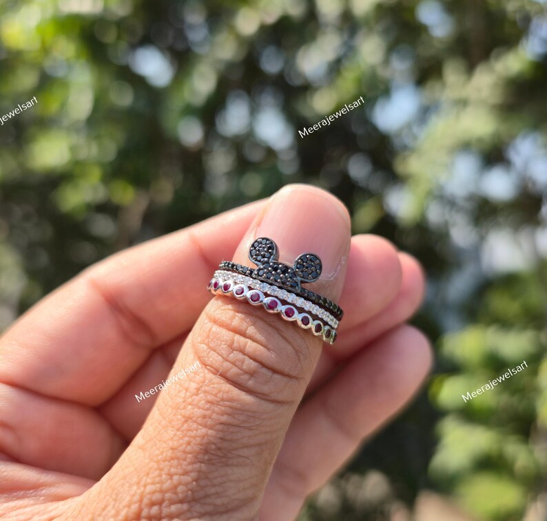 K&ouml;nnte beinhalten: Ein stapelbares Ring-Set bestehend aus einem oberen Ring mit einer mit schwarzen Steinen besetzten Mausohr-Silhouette, einem mittleren Ring mit klaren Pav&eacute;-Steinen und einem unteren Ring mit einer Reihe runder roter Steine.