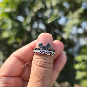 K&ouml;nnte beinhalten: Ein Stapel aus drei Ringen, bestehend aus einem oberen Ring mit einer mit schwarzen Steinen besetzten Mausohren-Silhouette, einem mittleren Ring mit einer Reihe klarer Pav&eacute;-Steine und einem unteren Ring mit runden roten Steinen in Fassung.