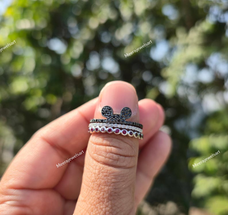 K&ouml;nnte beinhalten: Ein Stapel aus drei Ringen, bestehend aus einem oberen Ring mit einer mit schwarzen Steinen besetzten Mausohren-Silhouette, einem mittleren Ring mit einer Reihe klarer Pav&eacute;-Steine und einem unteren Ring mit runden roten Steinen in Fassung.