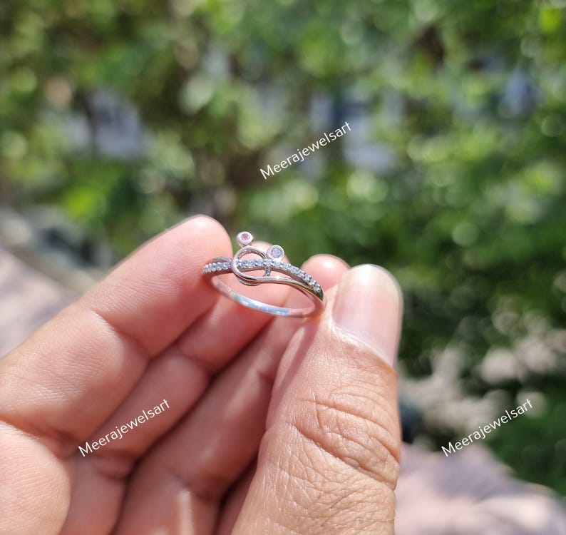 May include: A silver ring with a unique design, featuring small clear stones and a central knot-like detail. The ring is held in a hand, showcasing its delicate craftsmanship. The ring is a piece of jewelry.