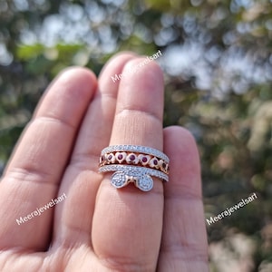 May include: A multi-band ring featuring a rose gold band with a row of small red heart-shaped stones and a silver-toned band with a bow-shaped accent, both embellished with tiny clear stones. The ring is worn on a finger with the text MeeraJewelsart.