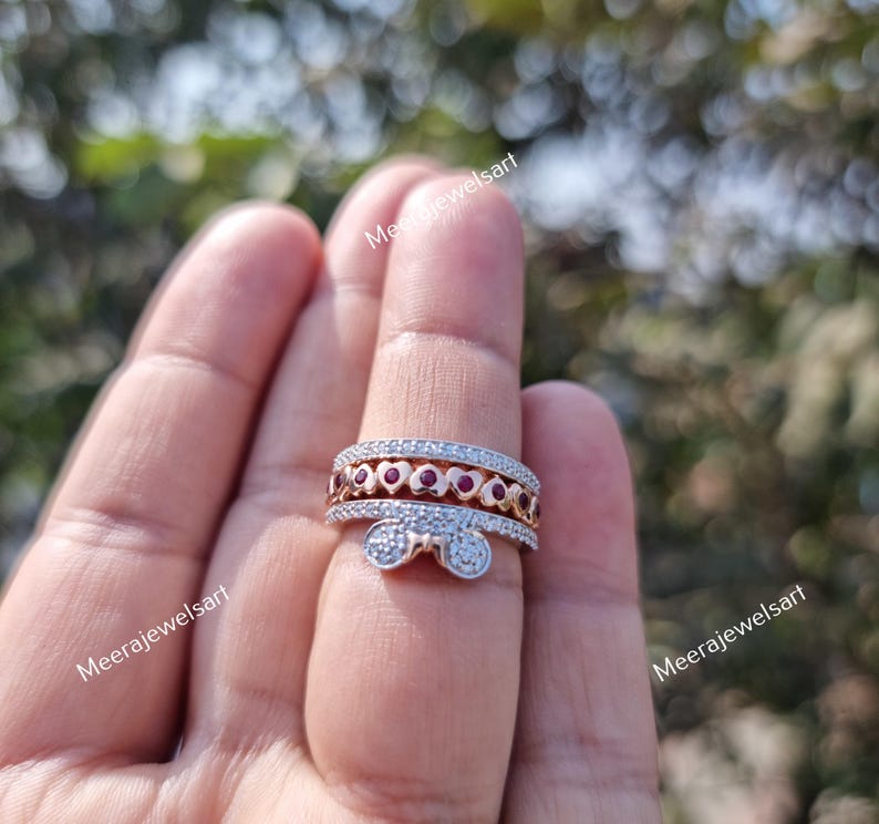May include: A multi-band ring featuring a rose gold band with a row of small red heart-shaped stones and a silver-toned band with a bow-shaped accent, both embellished with tiny clear stones. The ring is worn on a finger with the text MeeraJewelsart.