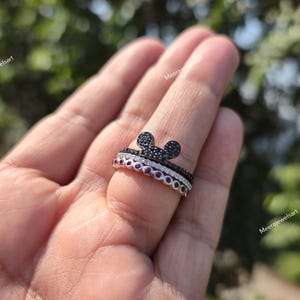 K&ouml;nnte beinhalten: Ein stapelbares Ring-Set mit einem oberen Ring, der eine mit schwarzen Steinen besetzte Mausohr-Silhouette zeigt, einem mittleren Ring mit klaren Steinen und einem unteren Ring mit einer Reihe runder roter Steine. Der Text Meerajewelsart ist sichtbar.