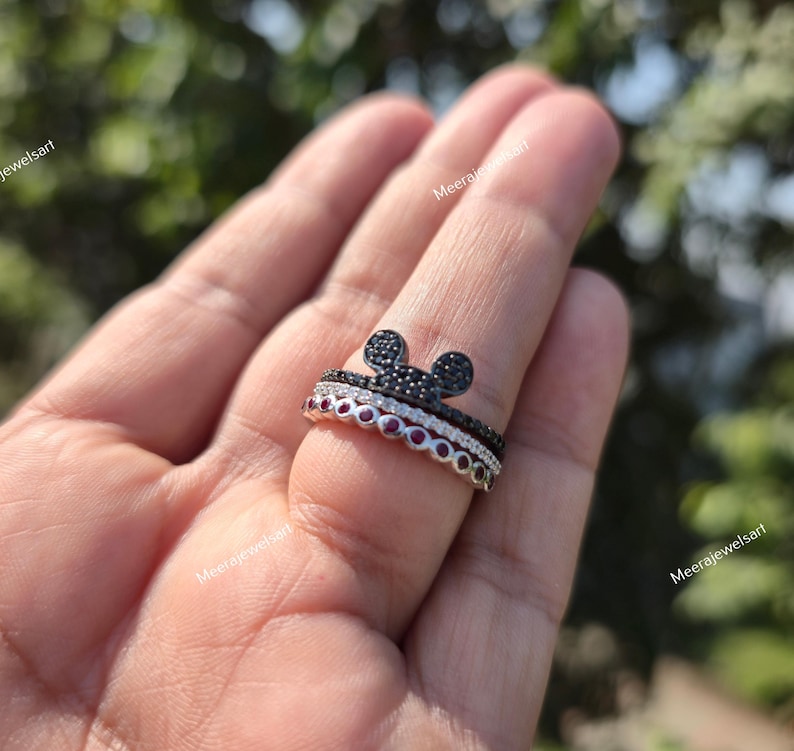 K&ouml;nnte beinhalten: Ein stapelbares Ring-Set mit einem oberen Ring, der eine mit schwarzen Steinen besetzte Mausohr-Silhouette zeigt, einem mittleren Ring mit klaren Steinen und einem unteren Ring mit einer Reihe runder roter Steine. Der Text Meerajewelsart ist sichtbar.