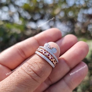 May include: A multi-band ring featuring a rose gold tone finish with a top band shaped like a mouse head silhouette encrusted with clear stones. The middle band displays a row of red heart-shaped stones, and the bottom band is a pav&eacute;-set clear stone band.