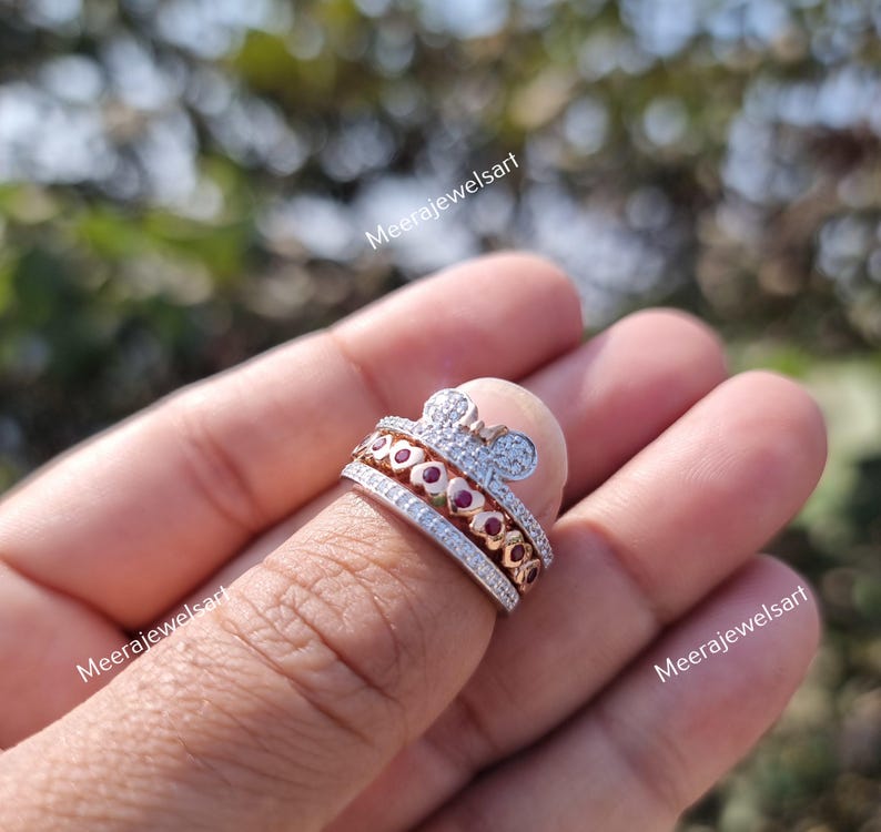May include: A multi-band ring featuring a rose gold tone finish with a top band shaped like a mouse head silhouette encrusted with clear stones. The middle band displays a row of red heart-shaped stones, and the bottom band is a pav&eacute;-set clear stone band.
