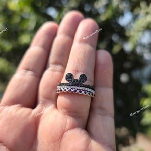 K&ouml;nnte beinhalten: Ein stapelbares Ring-Set bestehend aus drei Ringen. Der oberste Ring ist schwarz mit einer Mausohren-Silhouette, der mittlere Ring ist eine d&uuml;nne Reihe klarer Steine und der untere Ring besteht aus einer Reihe runder roter Steine in Silber.