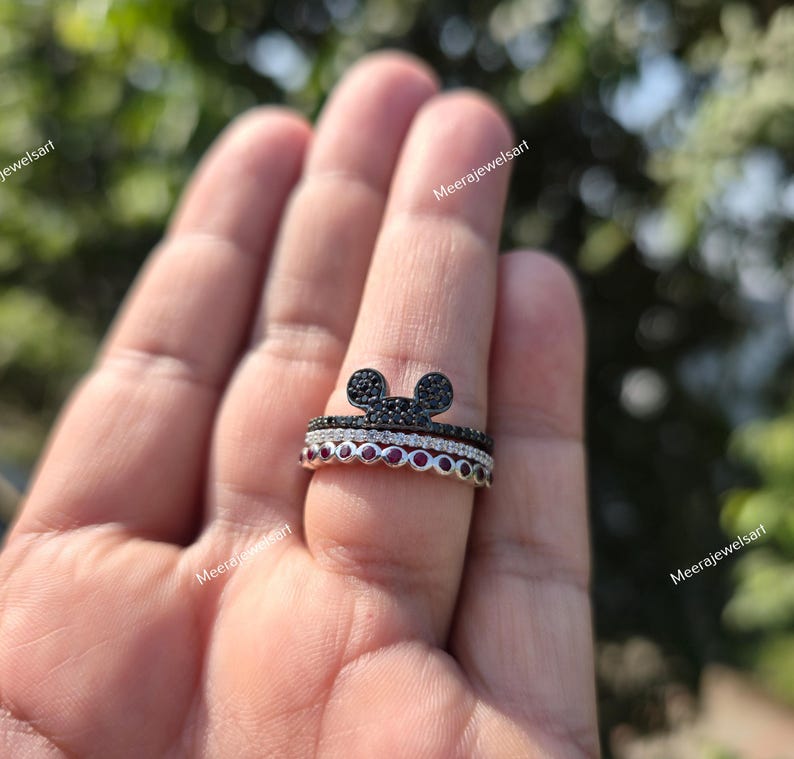 K&ouml;nnte beinhalten: Ein stapelbares Ring-Set bestehend aus drei Ringen. Der oberste Ring ist schwarz mit einer Mausohren-Silhouette, der mittlere Ring ist eine d&uuml;nne Reihe klarer Steine und der untere Ring besteht aus einer Reihe runder roter Steine in Silber.