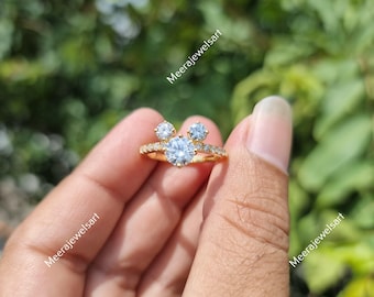 May include: A gold-colored ring with a large, round, clear gemstone in the center, flanked by two smaller, clear gemstones. The band is adorned with small, clear accent stones. The ring is held up against a blurred green background.
