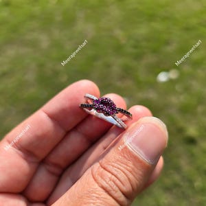 May include: A silver ring featuring a central cluster of small, round, deep pink stones surrounded by a halo of tiny black stones. The band is split into two thin, silver-toned arms that converge at the base, with the text Meerajewelsart visible.