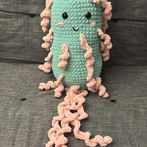 May include: A crocheted plush toy shaped like a bacteria with a smiling face. The toy is green with pink tentacles.
