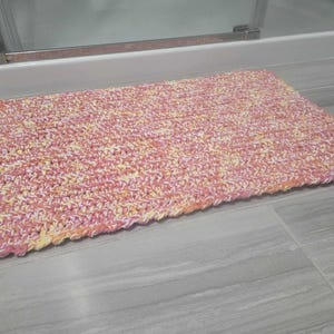 May include: A pink, yellow and white crocheted bath mat. The mat is made of soft yarn and has a textured surface.