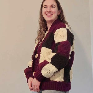 Peut inclure: Un cardigan en crochet avec un motif à carreaux en bordeaux, crème et noir. Le cardigan a une fermeture boutonnée et des manches longues.
