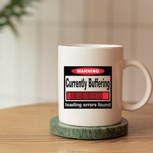 K&ouml;nnte beinhalten: Ein wei&szlig;er Keramikbecher mit einem rechteckigen Aufdruck, der ein rotes Oval mit dem Wort WARNING, den schwarzen Schriftzug Currently Buffering, einen roten Ladebalken sowie den Text loading errors found am unteren Rand enth&auml;lt.