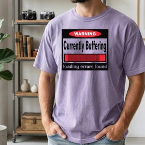 K&ouml;nnte beinhalten: Ein hellviolettes Kurzarm-T-Shirt mit einer schwarzen rechteckigen Grafik, die ein rotes Oval mit dem Wort WARNING, den Text Currently Buffering, einen roten Ladebalken und den Text loading errors found am unteren Rand enth&auml;lt.