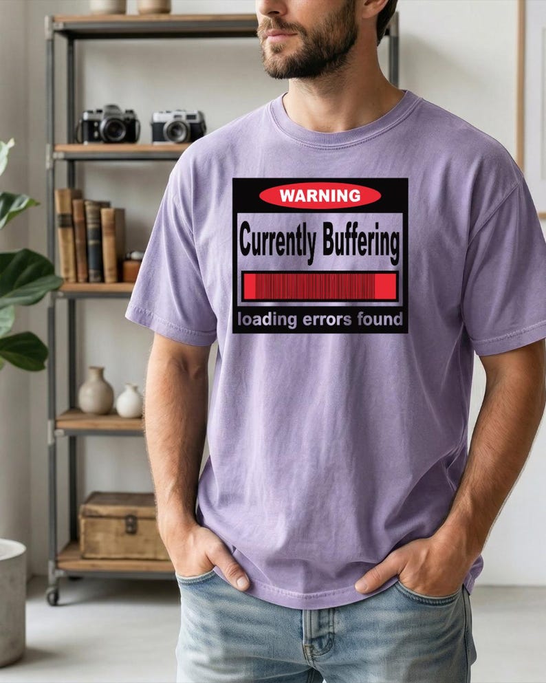 K&ouml;nnte beinhalten: Ein hellviolettes Kurzarm-T-Shirt mit einer schwarzen rechteckigen Grafik, die ein rotes Oval mit dem Wort WARNING, den Text Currently Buffering, einen roten Ladebalken und den Text loading errors found am unteren Rand enth&auml;lt.