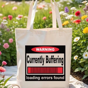 K&ouml;nnte beinhalten: Eine cremefarbene Stofftasche mit einer rechteckigen Grafik, die ein rotes Oval mit dem Wort WARNING, den Text Currently Buffering, einen roten Ladebalken und den Text loading errors found am unteren Rand enth&auml;lt.