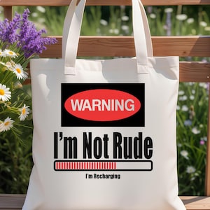 May include: A white tote bag featuring a black rectangular graphic with a red oval containing the word WARNING. Below this, the text reads I'm Not Rude followed by a loading bar graphic and the smaller text I'm Recharging in black lettering.