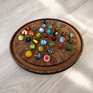 May include: A wooden, circular game board with a floral design, holding a collection of colorful glass marbles. The marbles are various colors and patterns, including blue, yellow, green, and red. The board is made of dark brown wood.