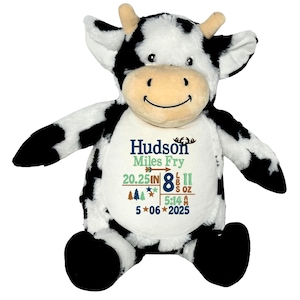 Peut inclure: Une peluche vache noire et blanche avec un visage souriant. La vache a un design personnalisé sur son ventre avec le nom "Hudson Miles Fry", des mesures et la date du 06/05/2025.