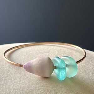 May include: A gold-toned bangle bracelet featuring three decorative beads. The beads include a light purple and white shell-shaped bead, and two sea glass beads in shades of turquoise and seafoam green. The bracelet is displayed on a neutral surface.