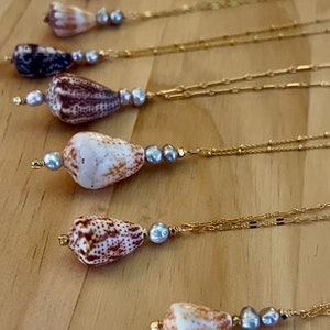 May include: Several gold necklaces with natural seashells and gray pearls. The seashells are various colors, including white, brown, and purple. The necklaces are displayed on a wooden surface.