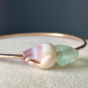 May include: A delicate gold-toned bracelet featuring two unique charms. One charm is a seashell with pink and white hues, and the other is a translucent, seafoam green glass bead. The bracelet is set against a soft, neutral background.