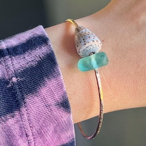 Puede incluir: Una pulsera dorada martillada con una concha y una cuenta de vidrio turquesa. La pulsera se lleva en la muñeca, con una manga con estampado tie-dye en el fondo. La concha tiene un patrón azul claro y marrón.