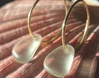 May include: A pair of gold hoop earrings with teardrop-shaped, sea glass pendants. The earrings are displayed on a pink seashell, with the gold hoops catching the light. The sea glass is a pale, translucent green.
