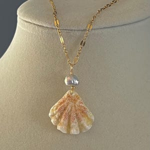 May include: A gold-toned necklace featuring a seashell pendant with a pearl accent. The seashell is cream-colored with pink and yellow hues. The necklace is displayed on a mannequin.